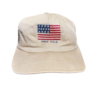 May include: A beige baseball cap featuring an embroidered American flag on the front panel. Below the flag, the text POLO U.S.A. is stitched in blue thread. The cap has a structured crown, a curved brim, and tonal stitching throughout the fabric.