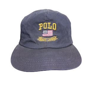 Pode incluir: Um boné de beisebol azul-marinho com o texto amarelo bordado POLO acima de um pequeno gráfico da bandeira americana. Abaixo da bandeira, há uma faixa amarela com o texto bordado RALPH LAUREN. O boné possui uma aba curva e detalhes costurados.
