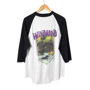 May include: A white baseball t-shirt with black three-quarter length sleeves featuring a graphic print on the front. The print displays the word WINDHAND in purple letters above a landscape scene of a river and trees with a dark shadow shape.
