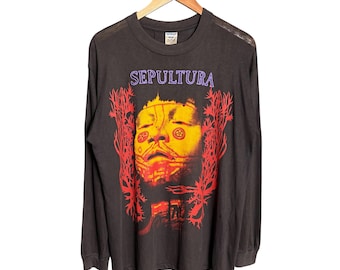 Puede incluir: Una camiseta de manga larga gris carbón con un estampado gráfico de un rostro en tonos amarillos y rojos. La palabra SEPULTURA está impresa en letras moradas sobre el rostro, con diseños rojos abstractos en los laterales.