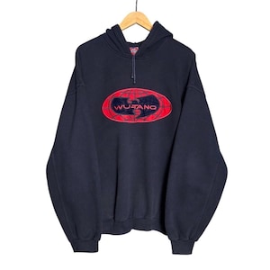 Può includere: Una felpa nera con cappuccio e maniche lunghe caratterizzata da una grande grafica ovale rossa sul petto. All'interno dell'ovale rosso si trova un logo Wu-Tang nero con un motivo a griglia. La felpa include un cordino e polsini a coste.