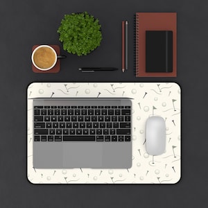 May include: A rectangular desk mat featuring a repeating pattern of golf balls, tees, and flags on a cream background. A silver laptop is centred on the mat with a white computer mouse to the right. The mat is placed on a dark grey surface.