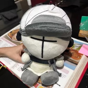 May include: A plush toy character with a white head, gray hat, and black cross-shaped face. The toy has gray arms, legs, and a gray outfit. The toy is sitting on top of a cookbook.