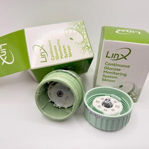 May include: A continuous glucose monitoring system with two light green components and two white and green boxes. The boxes have the "Linx" logo and text that reads "Continuous Glucose Monitoring System Sensor".
