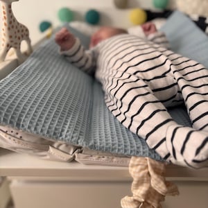 May include: A baby wearing a black and white striped onesie lies on a light blue waffle-textured changing pad. A giraffe toy stands nearby. Decorative pom-poms in various colors are in the background. A beige and white patterned scrunchie hangs from the changing table.