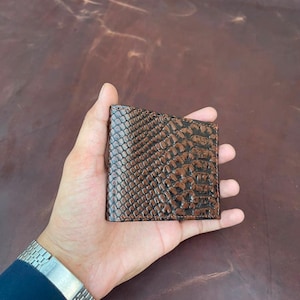 May include: A bi-fold wallet featuring a dark brown, textured exterior with a pattern resembling reptile scales. The wallet has a rectangular shape with visible stitching along the edges and a glossy finish that highlights the embossed texture.