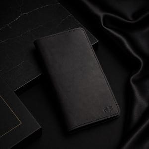 Könnte beinhalten: Ein rechteckiges schwarzes Brieftaschen-Etui mit glatter, matter Oberfläche und sichtbaren Nähten entlang der Kanten. In der unteren rechten Ecke befindet sich ein kleines geprägtes Logo aus zwei stilisierten, übereinanderliegenden F-Buchstaben.