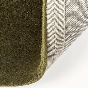 May include: Close-up of a section of a green rug with a textured surface. The rug's edge is visible, showing a contrasting beige and gray backing with a herringbone pattern. The rug is a solid olive green color.