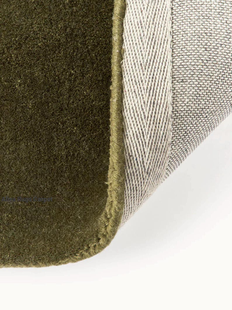 May include: Close-up of a section of a green rug with a textured surface. The rug's edge is visible, showing a contrasting beige and gray backing with a herringbone pattern. The rug is a solid olive green color.