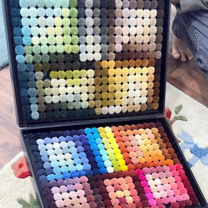 May include: A black case open to reveal a carpet color sample display. The display features numerous small, round carpet samples in a wide array of colors, including greens, blues, yellows, oranges, and pinks. The samples are arranged in a grid-like pattern, showcasing the variety of available carpet colors.
