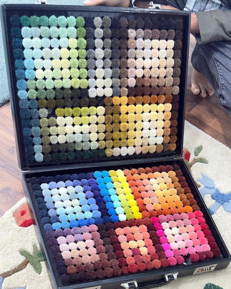 May include: A black case open to reveal a carpet color sample display. The display features numerous small, round carpet samples in a wide array of colors, including greens, blues, yellows, oranges, and pinks. The samples are arranged in a grid-like pattern, showcasing the variety of available carpet colors.