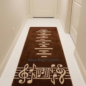 Puede incluir: Una alfombra rectangular marrón con un tema musical. La alfombra presenta una serie de líneas horizontales en el centro y notas musicales en la parte inferior. También se ve el texto "Aliya Rugs Carpet".