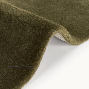 May include: Close-up of a section of a green rug with a soft, textured surface. The rug is folded over, revealing a glimpse of the backing. The rug's color is a muted olive green, and the texture appears plush.
