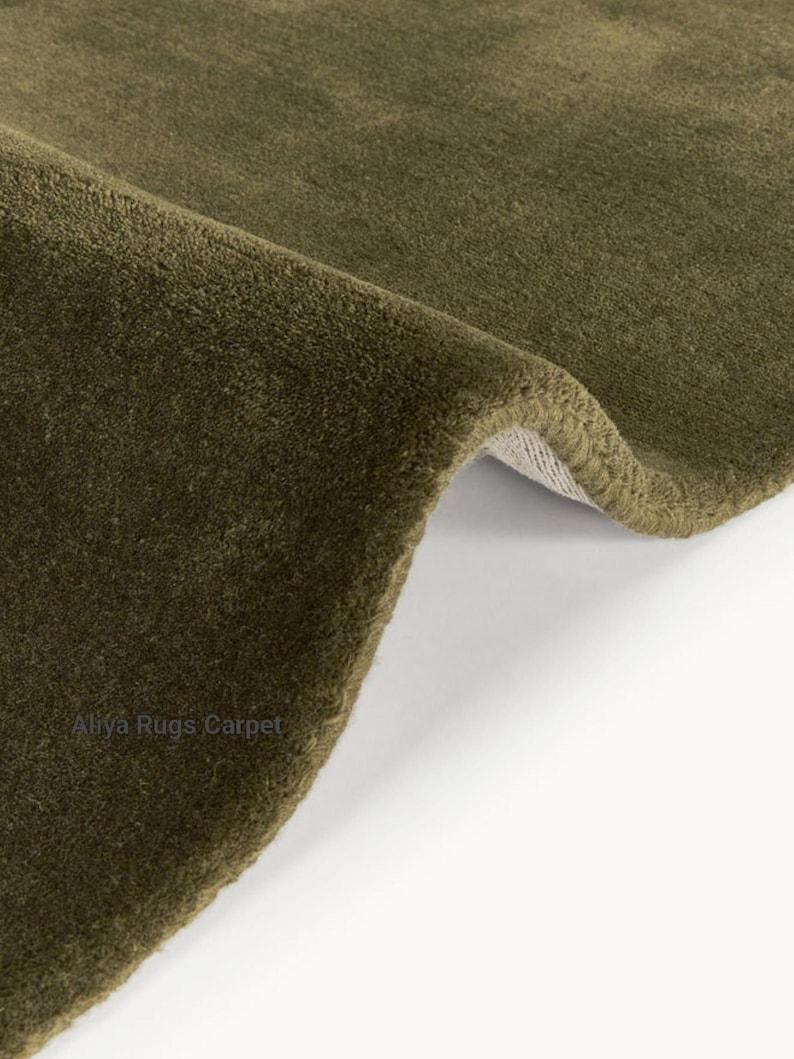 May include: Close-up of a section of a green rug with a soft, textured surface. The rug is folded over, revealing a glimpse of the backing. The rug's color is a muted olive green, and the texture appears plush.