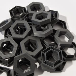 Puede incluir: Un montón de objetos hexagonales de color gris oscuro. Los objetos tienen un diseño geométrico con un centro hueco y una superficie texturizada. Los objetos están apilados sobre una superficie blanca.