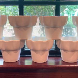 May include: A collection of nine white ceramic flower pots arranged in two rows. Each pot features a smooth, matte finish with a distinct scalloped, ruffled rim design. The pots are uniform in shape and size, displaying a simple, clean aesthetic.