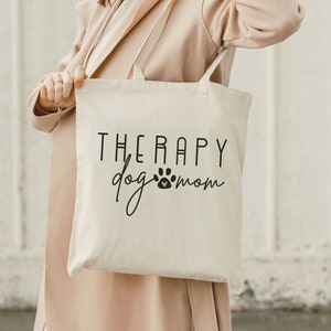 May include: A white canvas tote bag with black text that reads "Therapy dog mom" and a paw print with a pink heart.