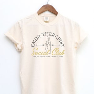 May include: A beige t-shirt with a graphic design of a hand making the "peace sign" gesture. The graphic is in a circle with the text "EMDR Therapist Social Club Going With That Since 1987".