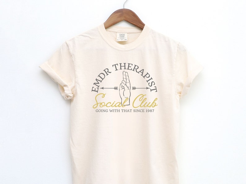 May include: A beige t-shirt with a graphic design of a hand making the "peace sign" gesture. The graphic is in a circle with the text "EMDR Therapist Social Club Going With That Since 1987".
