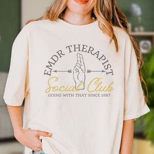 May include: A white t-shirt with a graphic design that says "EMDR Therapist Social Club Going With That Since 1987". The graphic features a hand making the peace sign with an arrow pointing to the right.