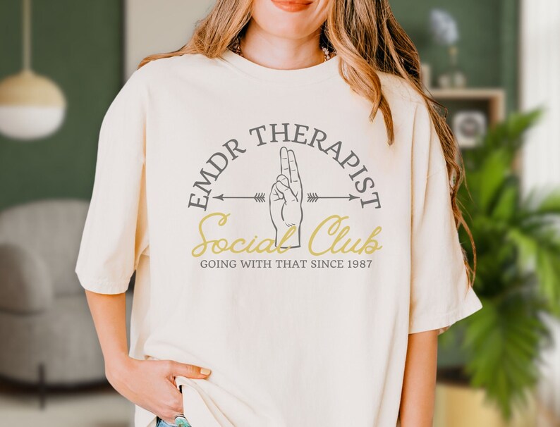 May include: A white t-shirt with a graphic design that says "EMDR Therapist Social Club Going With That Since 1987". The graphic features a hand making the peace sign with an arrow pointing to the right.