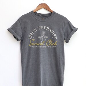 May include: Gray t-shirt with a white graphic design that says "EMDR Therapist Social Club Going With That Since 1987".