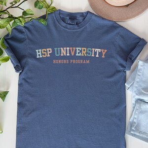 K&ouml;nnte beinhalten: Ein blaues T-Shirt mit dem Text "HSP UNIVERSITY HONORS PROGRAM" in bunten Buchstaben.