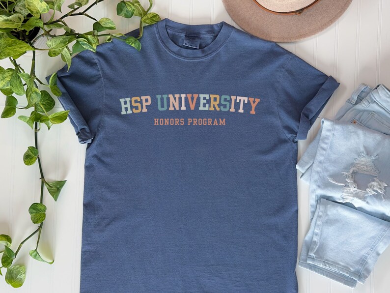 K&ouml;nnte beinhalten: Ein blaues T-Shirt mit dem Text "HSP UNIVERSITY HONORS PROGRAM" in bunten Buchstaben.