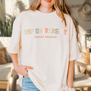 K&ouml;nnte beinhalten: Ein wei&szlig;es T-Shirt mit dem Text "HSP UNIVERSITY HONORS PROGRAM" in bunten Buchstaben.