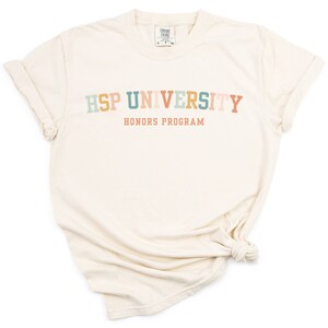 K&ouml;nnte beinhalten: Ein cremefarbenes T-Shirt mit dem Text "HSP UNIVERSITY HONORS PROGRAM" in Regenbogenfarben.
