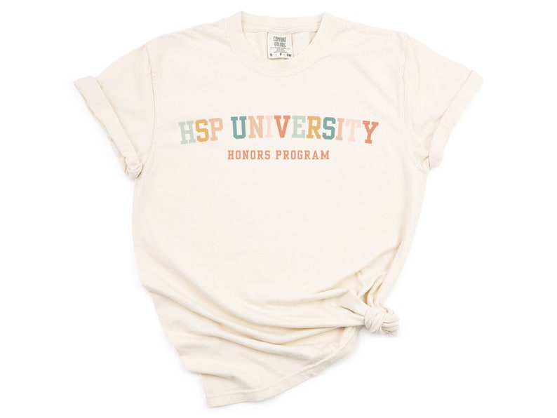 K&ouml;nnte beinhalten: Ein cremefarbenes T-Shirt mit dem Text "HSP UNIVERSITY HONORS PROGRAM" in Regenbogenfarben.