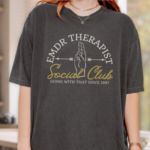 May include: A dark gray t-shirt with a white graphic design that says "EMDR Therapist Social Club Going With That Since 1987". The graphic includes a hand with two fingers raised and an arrow pointing to the right.