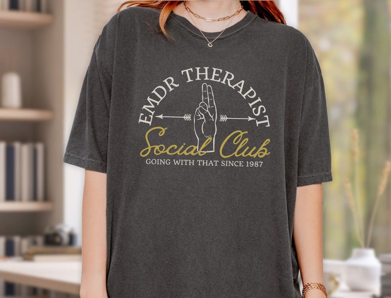 May include: A dark gray t-shirt with a white graphic design that says "EMDR Therapist Social Club Going With That Since 1987". The graphic includes a hand with two fingers raised and an arrow pointing to the right.