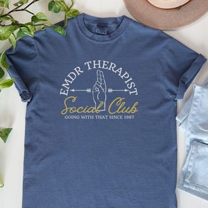 May include: A blue t-shirt with a white graphic design that says "EMDR Therapist Social Club Going With That Since 1987". The graphic also includes a hand making the peace sign and an arrow.