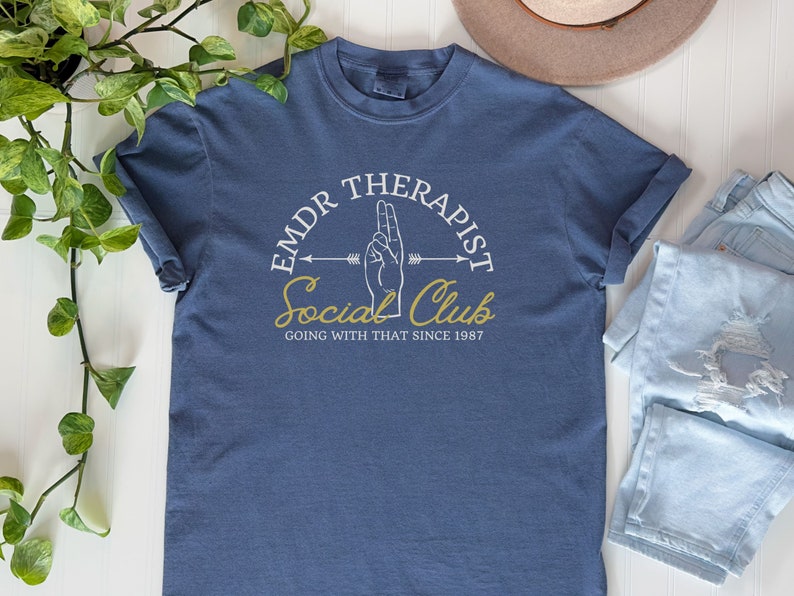 May include: A blue t-shirt with a white graphic design that says "EMDR Therapist Social Club Going With That Since 1987". The graphic also includes a hand making the peace sign and an arrow.