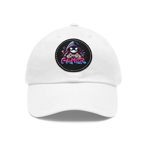 Könnte beinhalten: Weisse Baseballkappe mit gebogenem Schirm. Die Kappe hat einen runden, schwarzen Aufnäher mit einer farbenfrohen Grafik eines Kapuzencharakters, der Gamecontroller hält, und dem Wort "GAMER" in einer lebendigen Schriftart.