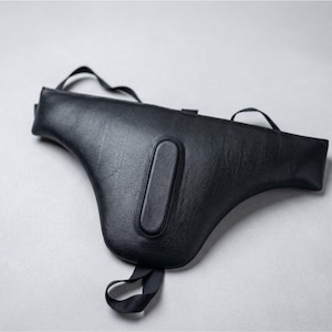 May include: A black leather holster with a curved shape and a rectangular accent. The holster has a strap and a handle. The product is designed to be worn on the body.