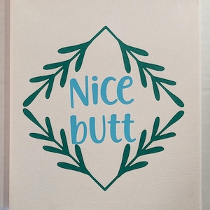 May include: Canvas art with the words "Nice Butt" in light blue, centered within a teal green leafy design. The background is a neutral off-white color.