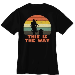 K&ouml;nnte beinhalten: Schwarzes T-Shirt mit Retro-Sonnenuntergangsdesign. Das Design zeigt eine Silhouette einer Figur mit Stab und einer kleinen Kreatur. Der orangefarbene Text darunter lautet "THIS IS THE WAY".