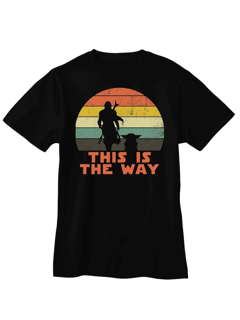 K&ouml;nnte beinhalten: Schwarzes T-Shirt mit Retro-Sonnenuntergangsdesign. Das Design zeigt eine Silhouette einer Figur mit Stab und einer kleinen Kreatur. Der orangefarbene Text darunter lautet "THIS IS THE WAY".