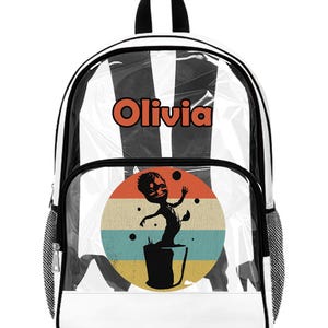 Könnte beinhalten: Weißer Rucksack mit schwarz-weißem Streifenmuster und transparentem Frontpanel. Er hat eine runde Grafik mit einer Cartoonfigur und dem Namen "Olivia" in Orange. Ideal für die Schule.