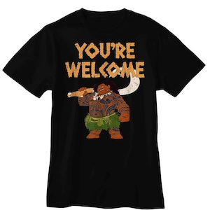 Puede incluir: Una camiseta negra de manga corta con un gr&aacute;fico de un personaje grande y musculoso con tatuajes tribales marrones, que lleva una falda de hojas verdes y sostiene un gran gancho blanco. El texto YOU'RE WELCOME est&aacute; impreso en naranja arriba.