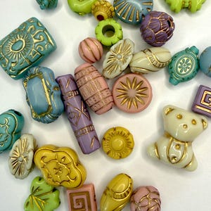 Op de afbeelding: Een verzameling kleurrijke kralen in verschillende vormen en maten. De kralen omvatten turquoise, geel, groen, roze en paars, met gouden accenten. Vormen zijn onder meer ovalen, vierkanten en een teddybeer. Ideaal voor het maken van sieraden.
