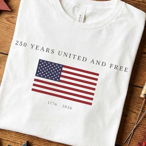 May include: A white t-shirt featuring a graphic of the United States flag centred on the chest. Above the flag, the text reads 250 YEARS UNITED AND FREE in black capital letters. Below the flag, the text reads 1776 - 2026 in black font.