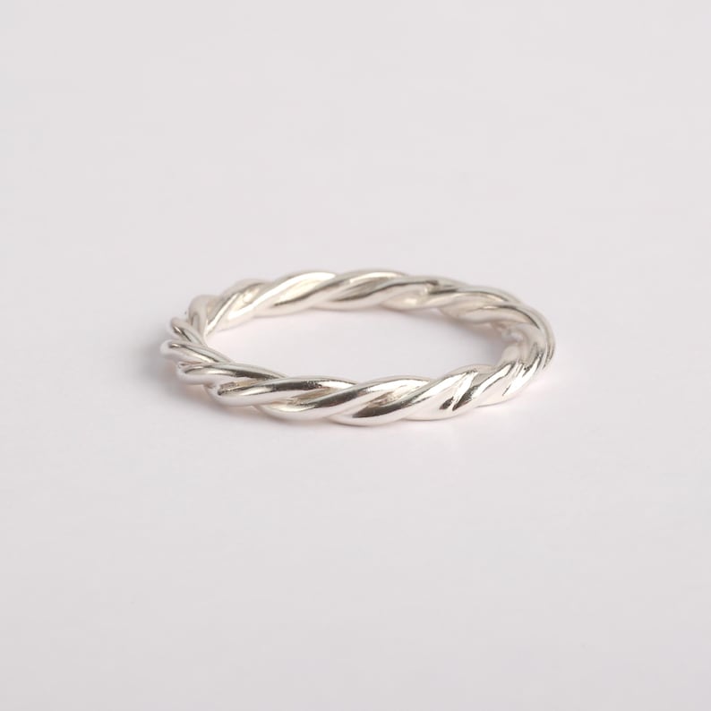 May include: A polished silver ring featuring a continuous twisted rope design. The band has a uniform metallic texture with reflective surfaces that catch the light, creating a series of subtle highlights and shadows along the circular metal structure.