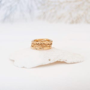 May include: A stack of three gold-toned rings featuring distinct textures. The top ring has a smooth surface, the middle ring displays a twisted rope design, and the bottom ring features a series of small, etched diagonal indentations.