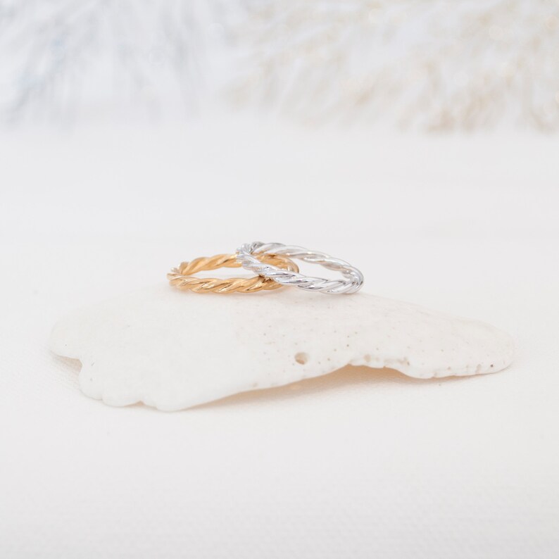 May include: Two twisted rings, one gold and one silver, are stacked on a white seashell. The rings are both simple and elegant.