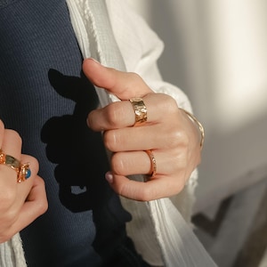 May include: A person's hand wearing three gold rings. The ring on the index finger is a wide band with a hammered texture. The ring on the middle finger is a thin band with a smooth finish. The ring on the ring finger is a thin band with a textured finish.
