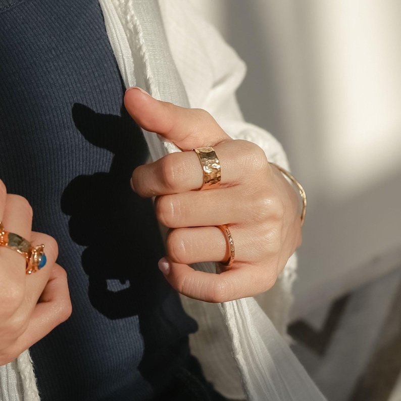 May include: A person's hand wearing three gold rings. The ring on the index finger is a wide band with a hammered texture. The ring on the middle finger is a thin band with a smooth finish. The ring on the ring finger is a thin band with a textured finish.