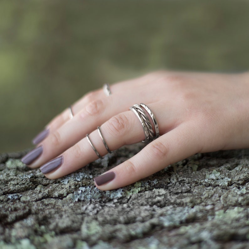 May include: A hand wearing multiple silver rings, including a twisted band ring, on a finger. The hand is resting on a textured, brown surface.